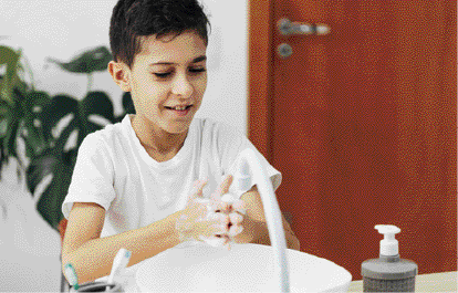 Imagem: Fotografia. Um menino com cabelo curto e camiseta branca está lavando as mãos sobre uma pia. Ao fundo, um vaso com planta e uma porta.  Fim da imagem.