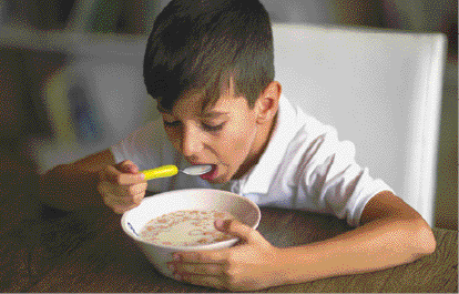 Imagem: Fotografia. O menino está sentado e segurando uma colher na frente da boca. Com a outra mão, ele segura uma tigela com cereal e leite.  Fim da imagem.