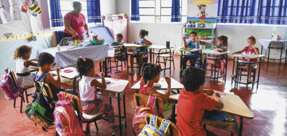 Imagem: Fotografia. No centro há duas fileiras de carteiras escolares com crianças sentadas. No centro, uma mulher está em pé, atrás de uma mesa grande com objetos em cima. Fim da imagem.