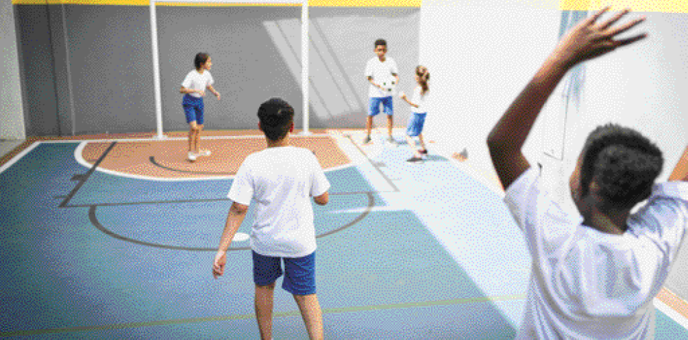 Imagem: Fotografia. Cinco crianças com camiseta branca e bermuda azul estão brincando com uma bola em uma quadra. Fim da imagem.