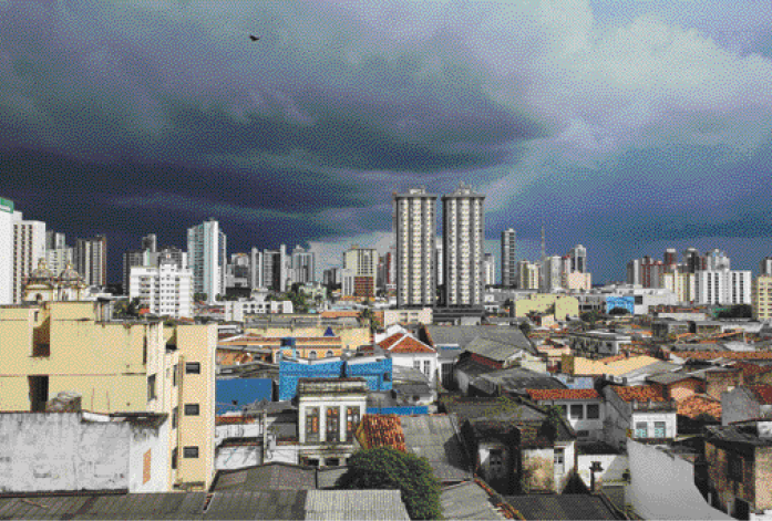 Imagem: Fotografia. Uma cidade com casas e prédios. Acima, nuvens escuras no céu.  Fim da imagem.