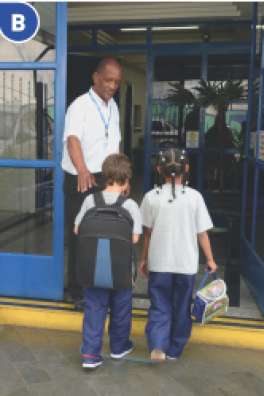 Imagem: Fotografia B. Duas crianças com camiseta branca, calça azul e tênis estão de costas e entrando em um prédio. Ao lado, um homem com camisa branca e calça preta sorri. Fim da imagem.