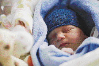 Imagem: Fotografia. Um recém-nascido com gorro azul está dormindo com um cobertor azul em volta do corpo. Fim da imagem.