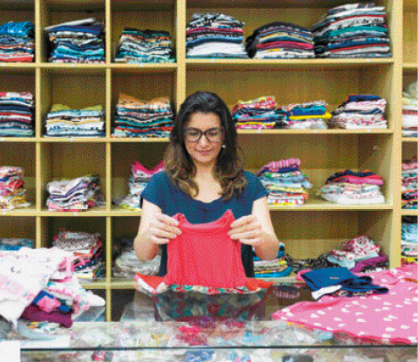 Imagem: Fotografia. Uma mulher está dobrando um vestido infantil sobre um balcão. Atrás dela há várias roupas dobradas em uma prateleira.  Fim da imagem.