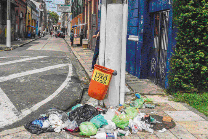 Imagem: Fotografia. Muito lixo em volta de um cesto de lixo, que está pendurado em um poste. Ao fundo, casas e carros. Fim da imagem.