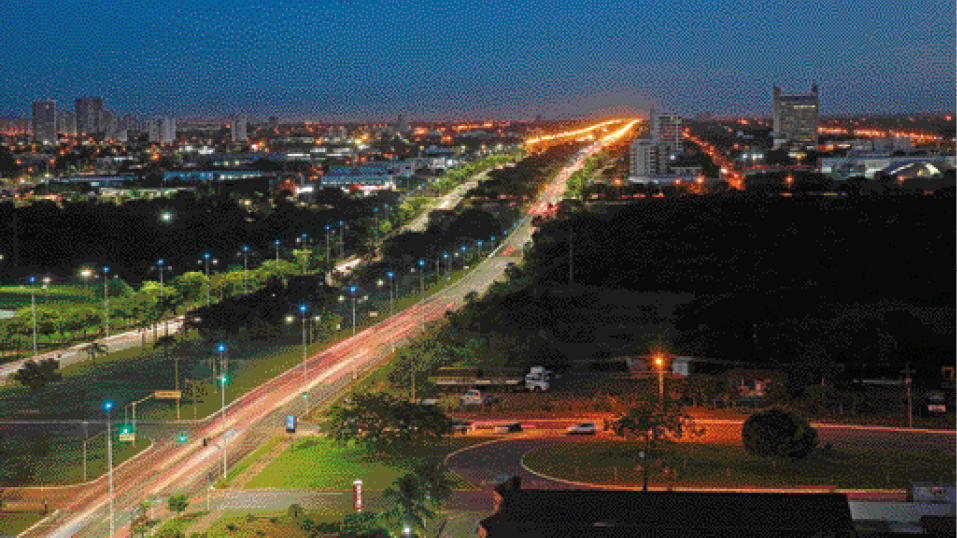 Imagem: Fotografia. Vista aérea de uma avenida vazia com postes acesos ao lado. Nas laterais há sombras de árvores e ao fundo, luzes acesas em prédios. Fim da imagem.