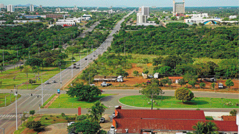 Imagem: Fotografia. Vista aérea de uma avenida com carros. Ao lado há árvores e ao fundo, prédios. Fim da imagem.