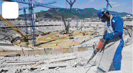 Imagem: Fotografia. Uma pessoa com capacete de proteção, máscara, avental e uniforme azul está segurando uma britadeira sobre uma construção.  Fim da imagem.