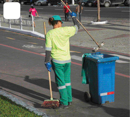 Imagem: Fotografia. Uma mulher com uniforme verde está segurando uma vassoura e uma pá e jogando lixo dentro de um cesto. Ao fundo, pessoas andando na calçada. Fim da imagem.