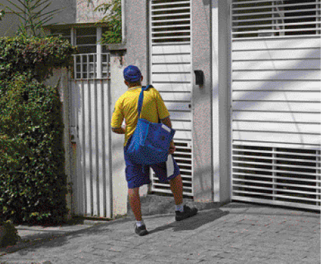 Imagem: Fotografia. Um homem com boné e bermuda azul e camiseta amarela está de costas e segurando uma bolsa grande no ombro direito. Na frente dele há uma casa e plantas. Fim da imagem.