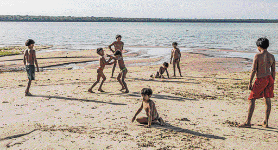 Imagem: Fotografia. Crianças indígenas com bermudas coloridas estão brincando na areia. Ao fundo, uma lagoa e o céu em tons de azul. Fim da imagem.