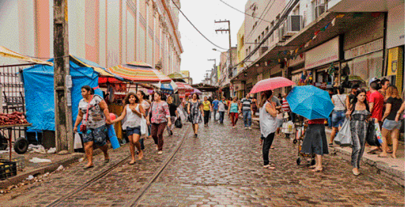 Imagem: Fotografia. Pessoas segurando guarda-chuvas em uma rua de pedras. Nas laterais há lojas e postes.  Fim da imagem.