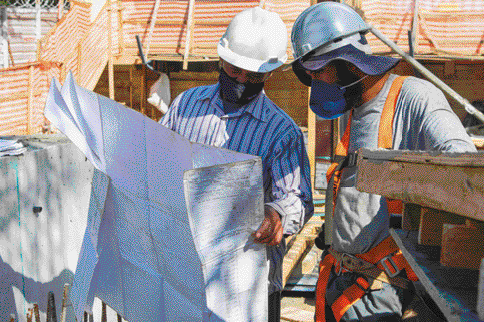 Imagem: Fotografia. Duas pessoas com capacete de proteção e máscara estão olhando um papel grande. Ao fundo, construção.  Fim da imagem.