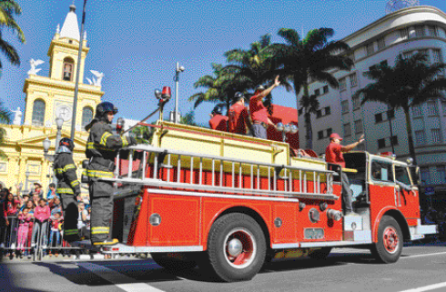 Imagem: Fotografia. Homens com uniforme vermelho estão acenando no topo de um caminhão de bombeiros. Em volta, pessoas observam e ao fundo há árvores e prédios. Fim da imagem.