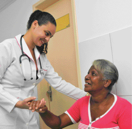 Imagem: Fotografia. Uma senhora está sentada e ao seu lado, uma mulher com jaleco branco e estetoscópio pendurado no pescoço está sorrindo e segurando sua mão.  Fim da imagem.