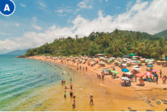 Imagem: Fotografia A. Uma multidão na areia da praia e entre as pessoas há guarda-sóis coloridos. Ao lado, o mar em tons de verde e ao fundo, árvores e morros. Fim da imagem.