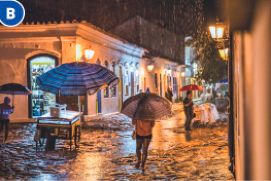 Imagem: Fotografia B. Pessoas andando em uma rua de pedras e segurando guarda-chuvas. Em volta delas há chuva caindo e ao fundo, construções com as luzes acesas. Fim da imagem.