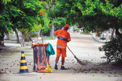 Imagem: Fotografia. Um homem com boné e uniforme laranja está varrendo uma rua. Ao seu lado há um cesto de lixo e um cone. Ao fundo, árvores. Fim da imagem.