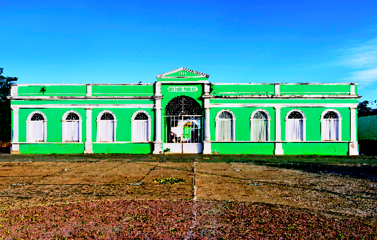 Imagem: Fotografia. Fachada de uma construção extensa com parede verde e detalhes brancos. Ao fundo, árvores e o céu azul.  Fim da imagem.