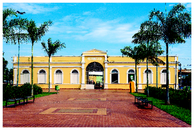 Imagem: Fotografia. Fachada de uma construção extensa com parede amarela e detalhes brancos. Na frente há um pátio com chão marrom, plantas, árvores e bancos. Ao fundo, árvores e o céu azul.  Fim da imagem.