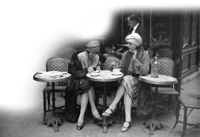 Imagem: Fotografia em preto e branco. Duas mulheres com chapéu, casaco e vestidos estão sentadas ao lado de uma mesa pequena e redonda. Ao fundo, um homem com terno e gravata está em pé e segurando uma bandeja. Fim da imagem.