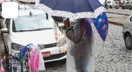 Imagem: Fotografia. Uma pessoa com capa de chuva transparente está segurando um guarda-chuva colorido sobre a cabeça. Ao seu lado há um carrinho com vários guarda-chuvas dentro. Ao fundo, carros em uma rua. Fim da imagem.