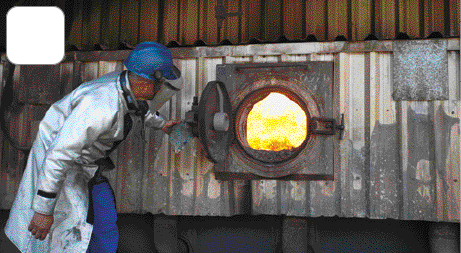 Imagem: Fotografia. Uma pessoa com capacete de proteção, máscara e jaleco está abrindo um forno grande com brasa dentro. Fim da imagem.