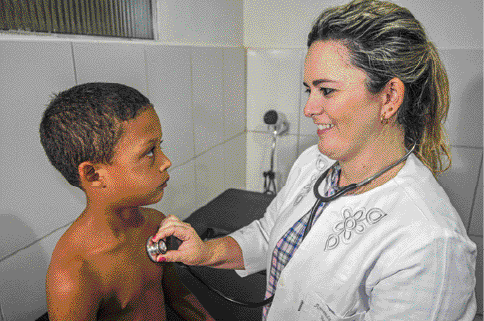 Imagem: Fotografia. Um menino com cabelo curto e sem camiseta está sentado. Na frente dele, uma mulher com cabelo preso e jaleco branco está com um estetoscópio pendurado no pescoço e segurando a ponta do objeto no peito do menino. Fim da imagem.