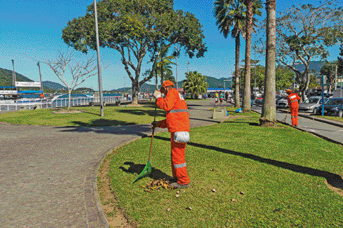 Imagem: Fotografia. Duas pessoas com uniforme laranja estão varrendo uma praça. Ao fundo, árvores e o mar. Fim da imagem.