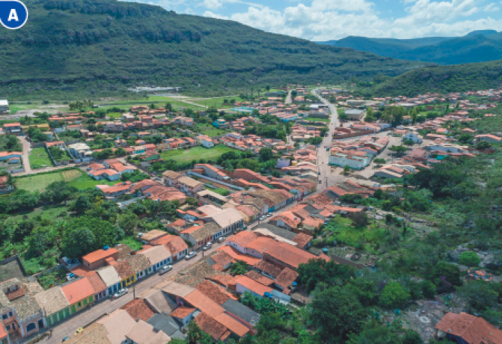 Imagem: Fotografia A. Vista aérea de uma cidade com casas, carros e árvores. Ao fundo, morros e nuvens no céu azul.  Fim da imagem.