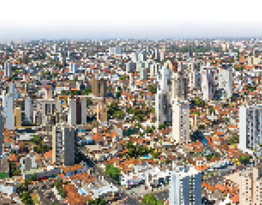 Imagem: Fotografia. Vista aérea de uma cidade com casas, prédios, ruas e árvores. Fim da imagem.