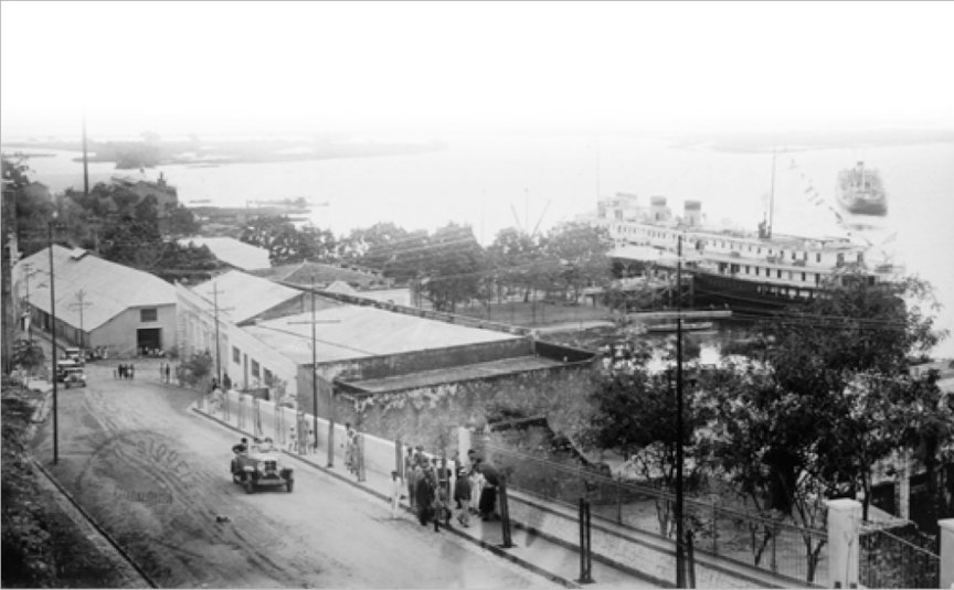 Imagem: Fotografia em preto e branco. À esquerda, um carro andando sobre uma rua de terra. À direita, pessoas na calçada e ao lado há construções e árvores. Ao fundo, navios no mar. Fim da imagem.