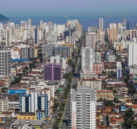 Imagem: Fotografia. Vista aérea de uma avenida asfaltada. Nas laterais há vários prédios, casas e algumas árvores. Fim da imagem.