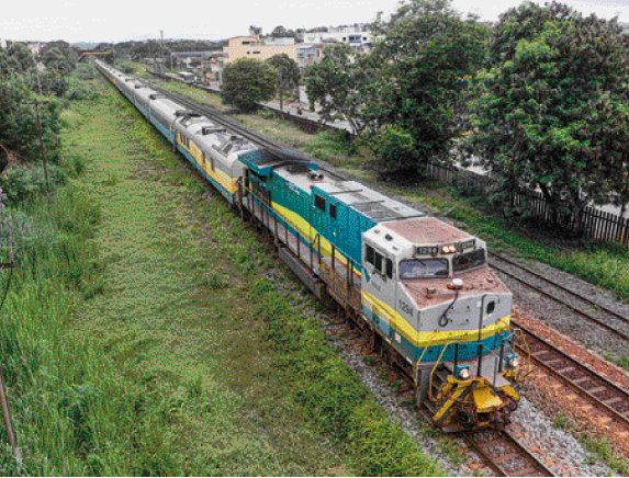 Imagem: Fotografia. Vista aérea de um trem verde e amarelo andando sobre trilhos. Nas laterais há árvores.  Fim da imagem.