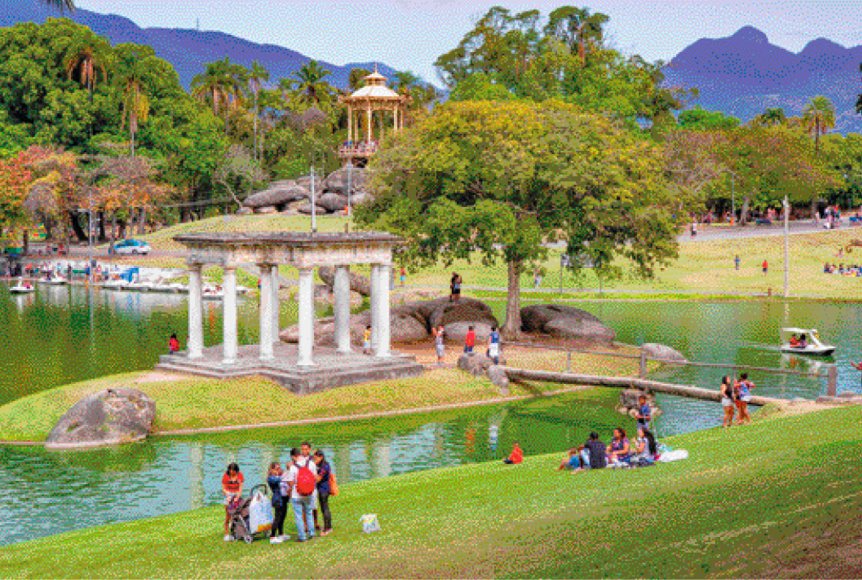 Imagem: Fotografia. No centro, famílias sentadas em um gramado ao lado de um lago com pedalinhos. Ao lado há uma ponte de madeira e do meio do lago há pedras, uma árvore e colunas grandes e brancas. Ao fundo, mais pessoas, árvores e morros. Fim da imagem.