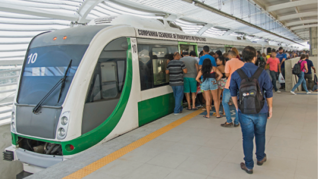 Imagem: Fotografia. Pessoas aglomeradas ao lado de um trem cinza e verde, que está parado em uma plataforma coberta. Fim da imagem.