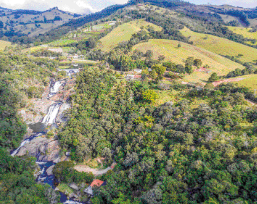 Imagem: Fotografia. Vista aérea de algumas casas espalhadas em uma serra com várias árvores. Ao fundo, mais serras.   Fim da imagem.