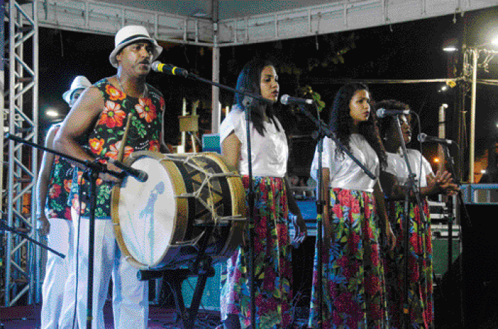 Imagem: Fotografia. Três mulheres com blusa branca e saia florida estão lado a lado e cantando atrás de microfones. Ao lado, um homem com chapéu branco, regata florida e calça branca está tocando tambor.  Fim da imagem.