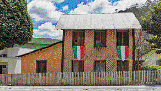Imagem: Fotografia. Casa com dois andares e paredes de barro. Nas janelas há duas bandeiras da Itália. Ao fundo, casas e árvores.  Fim da imagem.