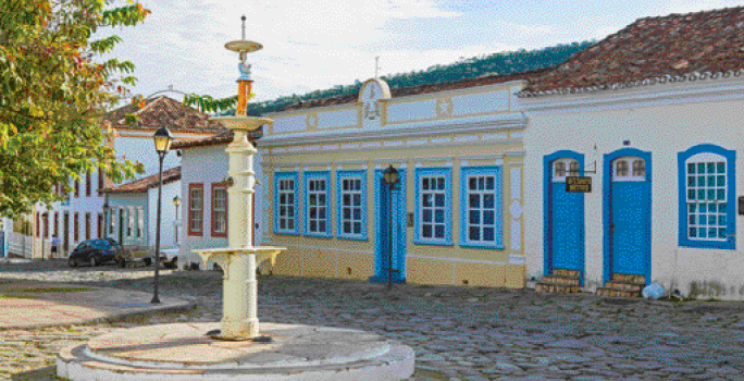 Imagem: Fotografia. Casas lado a lado com paredes em tons de bege e detalhes coloridos. Ao lado, rua de pedras e árvores. Fim da imagem.