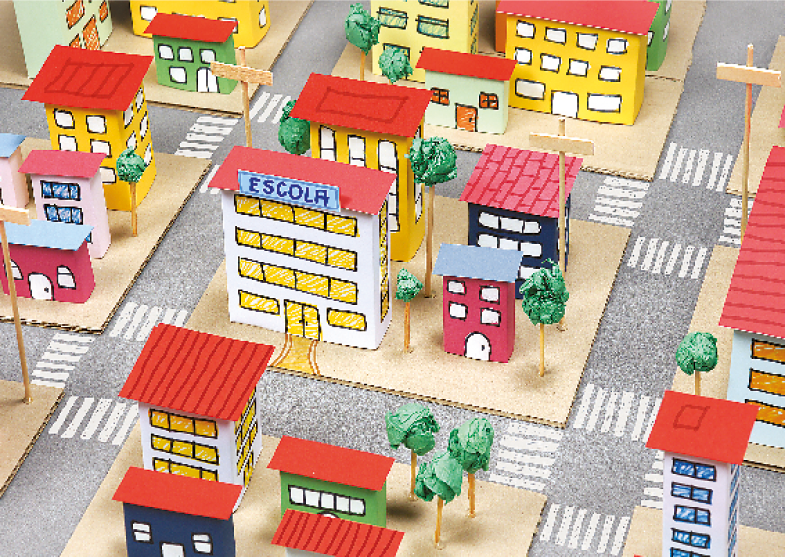 Imagem: Fotografia. Vista aérea de uma maquete de bairro. No centro, a escola e em volta, prédios, casas, árvores e ruas.  Fim da imagem.