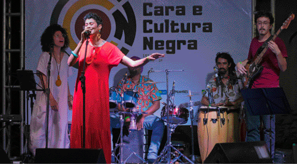 Imagem: Fotografia. No centro, uma mulher com vestido vermelho está cantando em um palco. Atrás dela há homens estão tocando instrumentos musicais e em seguida há um cartaz com a informação: CARA E CULTURA NEGRA. Fim da imagem.