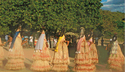 Imagem: Fotografia. Pessoas com roupa de palha estão dançando. Ao fundo, árvores.   Fim da imagem.