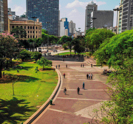 Imagem: Fotografia. Vista aérea de uma praça com árvores. À direita há pessoas andando em uma calçada larga em tons de marrom. Ao fundo, prédios.   Fim da imagem.