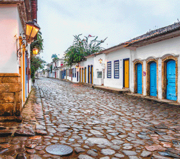 Imagem: Fotografia. Casas lado a lado com paredes brancas e detalhes coloridos. No centro há uma rua de pedras e em volta, árvores.   Fim da imagem.