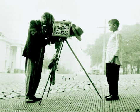Imagem: Fotografia em preto e branco. Um homem com terno está atrás de uma câmera fotográfica e com um tecido sobre a cabeça. Na frente dele, um menino está em pé e olhando para frente. Ao fundo, árvores.  Fim da imagem.