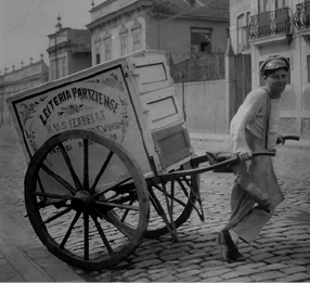 Imagem: Fotografia em preto e branco. Um jovem está sorrindo e puxando uma charrete e acima dela há uma caixa grande com a informação: LEITERIA. Ao fundo, construções.   Fim da imagem.