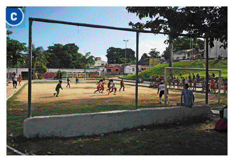 Imagem: Fotografia C. Pessoas jogando futebol em um campo de terra. Ao fundo, casas e árvores.   Fim da imagem.