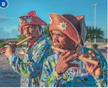 Imagem: Fotografia D. Dois homens com chapéus de cangaceiros e camisa florida estão segurando pífanos na frente da boca. Ao fundo, árvores.  Fim da imagem.