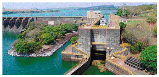 Imagem: Fotografia. Ao fundo, uma represa e na frente, uma barragem. Ao lado há uma eclusa com um navio parado.  Fim da imagem.